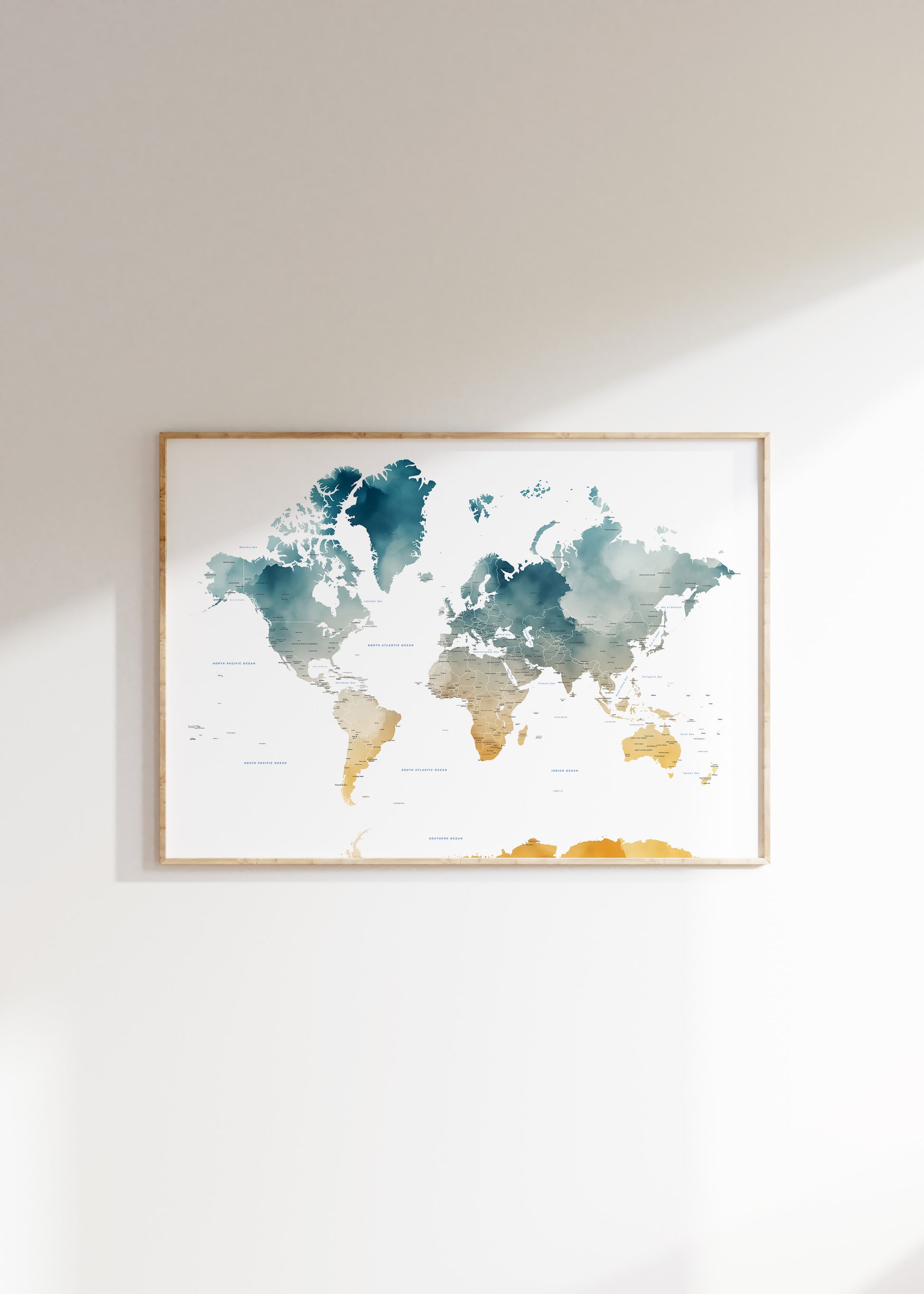 A framed world map watercolor print with teal, blue, and gold landmasses on a white background, hanging on a light beige wall with soft sunlight casting shadows.