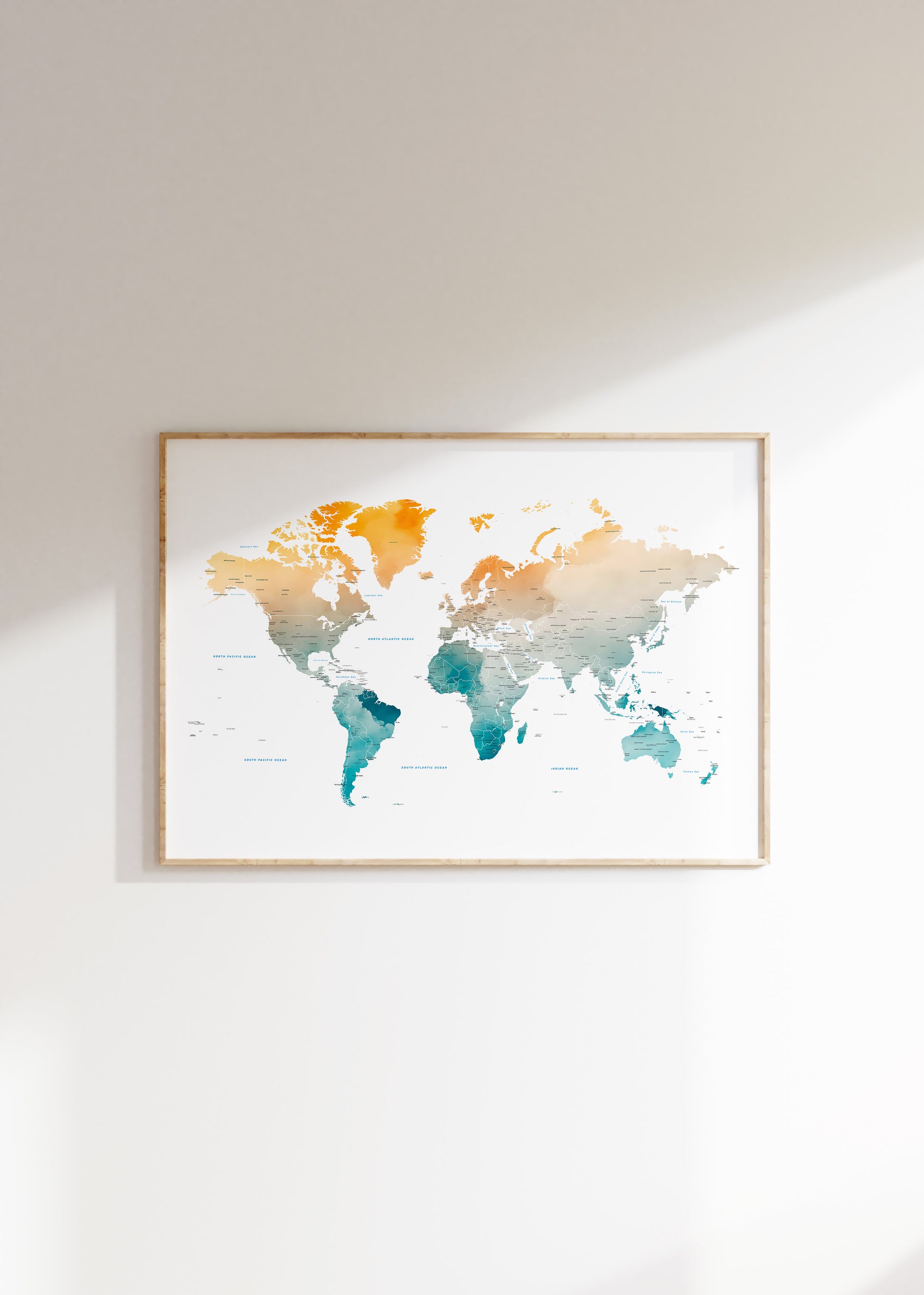 A framed world map watercolor print with a gradient of orange, beige, and teal blue, hanging on a white wall with soft sunlight casting a shadow.