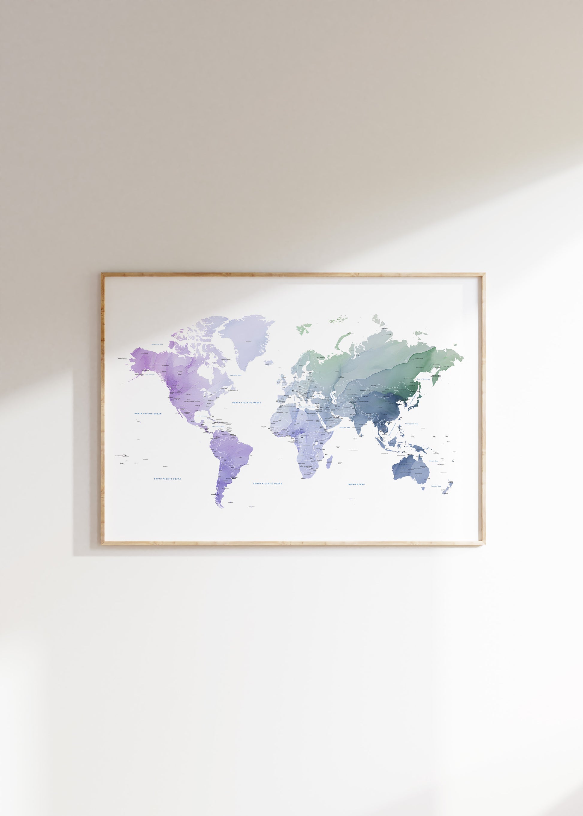 A framed world map watercolor print in shades of purple, blue, and green, hanging on a light beige wall with soft sunlight casting shadows.