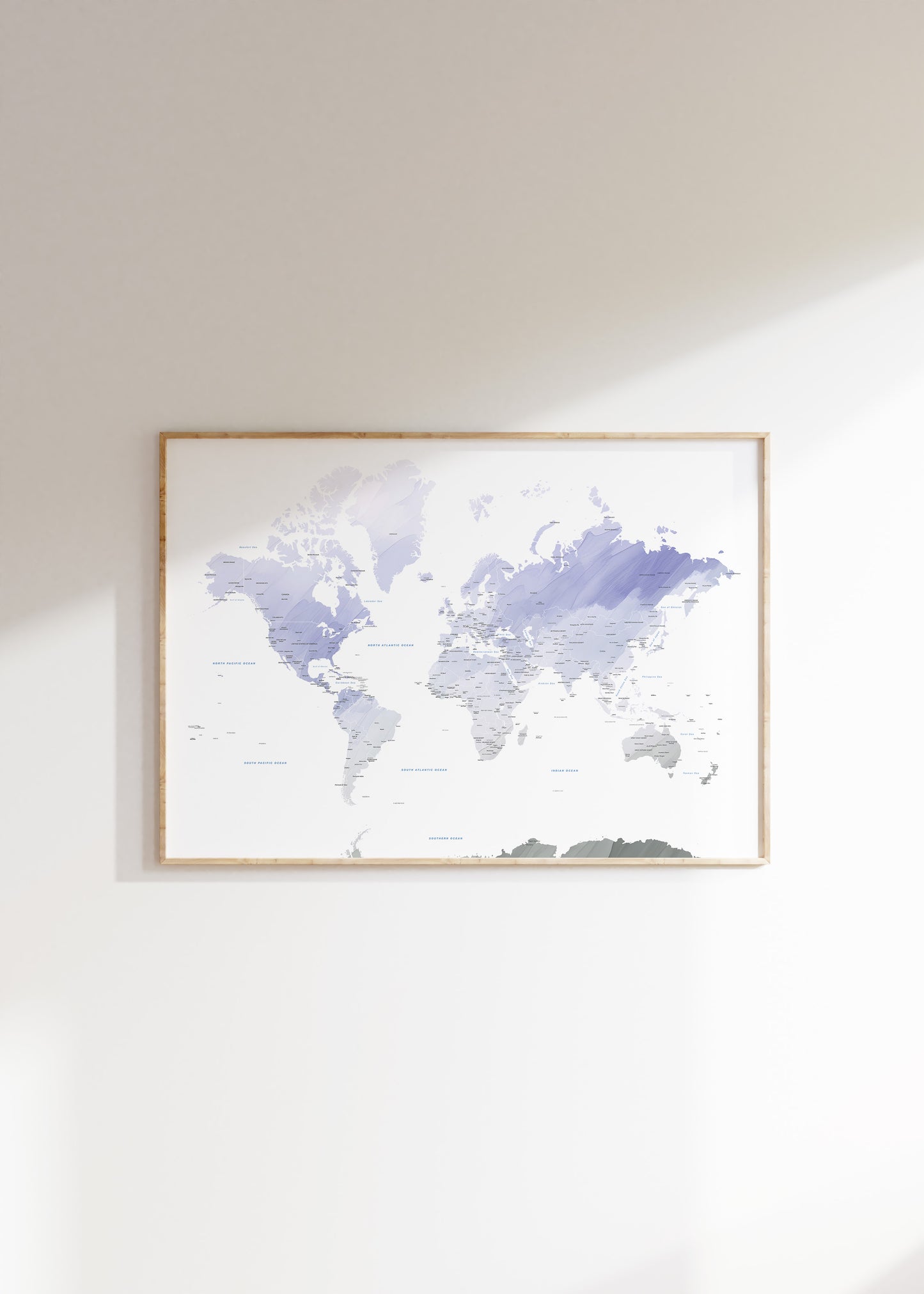 A framed world map art print with watercolor-style continents in shades of lavender, purple, and gray on a white background, hung on a light beige wall.