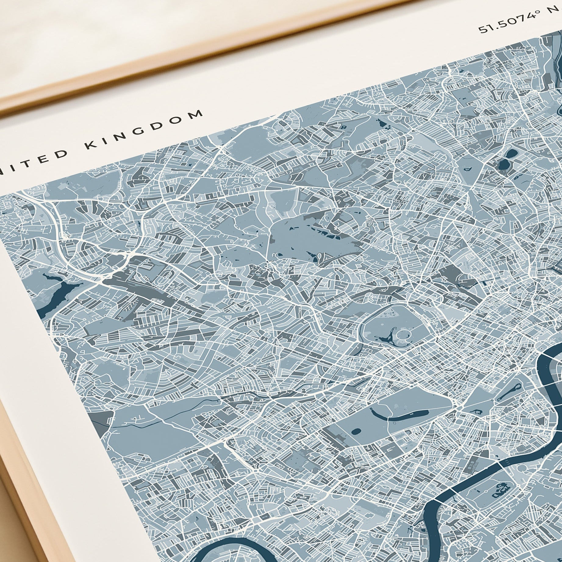 A framed London city map print with a light blue and white street layout, shown resting on a beige tabletop next to a cream gift box.