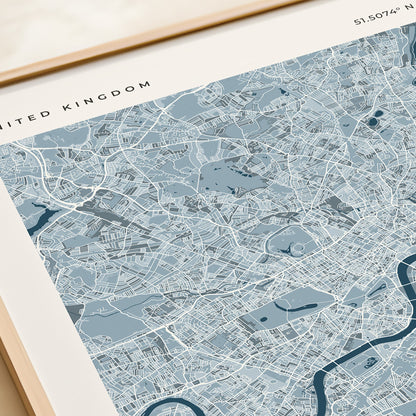A framed London city map print with a light blue and white street layout, shown resting on a beige tabletop next to a cream gift box.