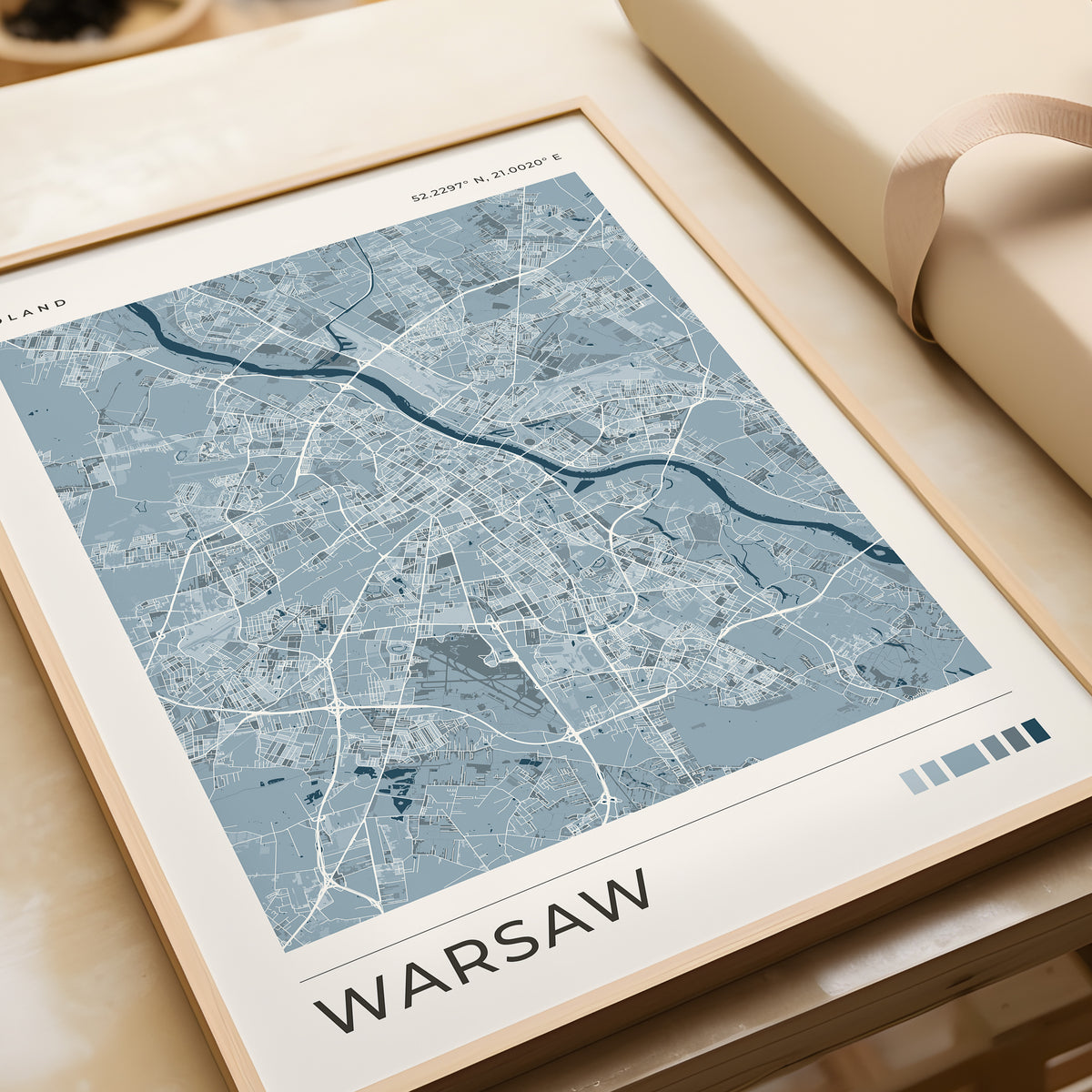 A framed city map print of Warsaw, Poland, featuring a light blue and white street layout with a cream border, styled on a beige tabletop next to a rolled cream paper or gift wrap.