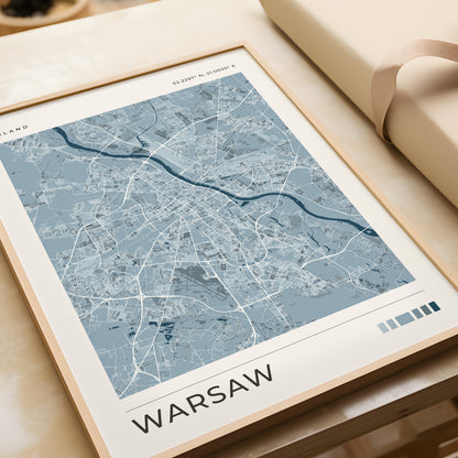 A framed city map print of Warsaw, Poland, featuring a light blue and white street layout with a cream border, styled on a beige tabletop next to a rolled cream paper or gift wrap.