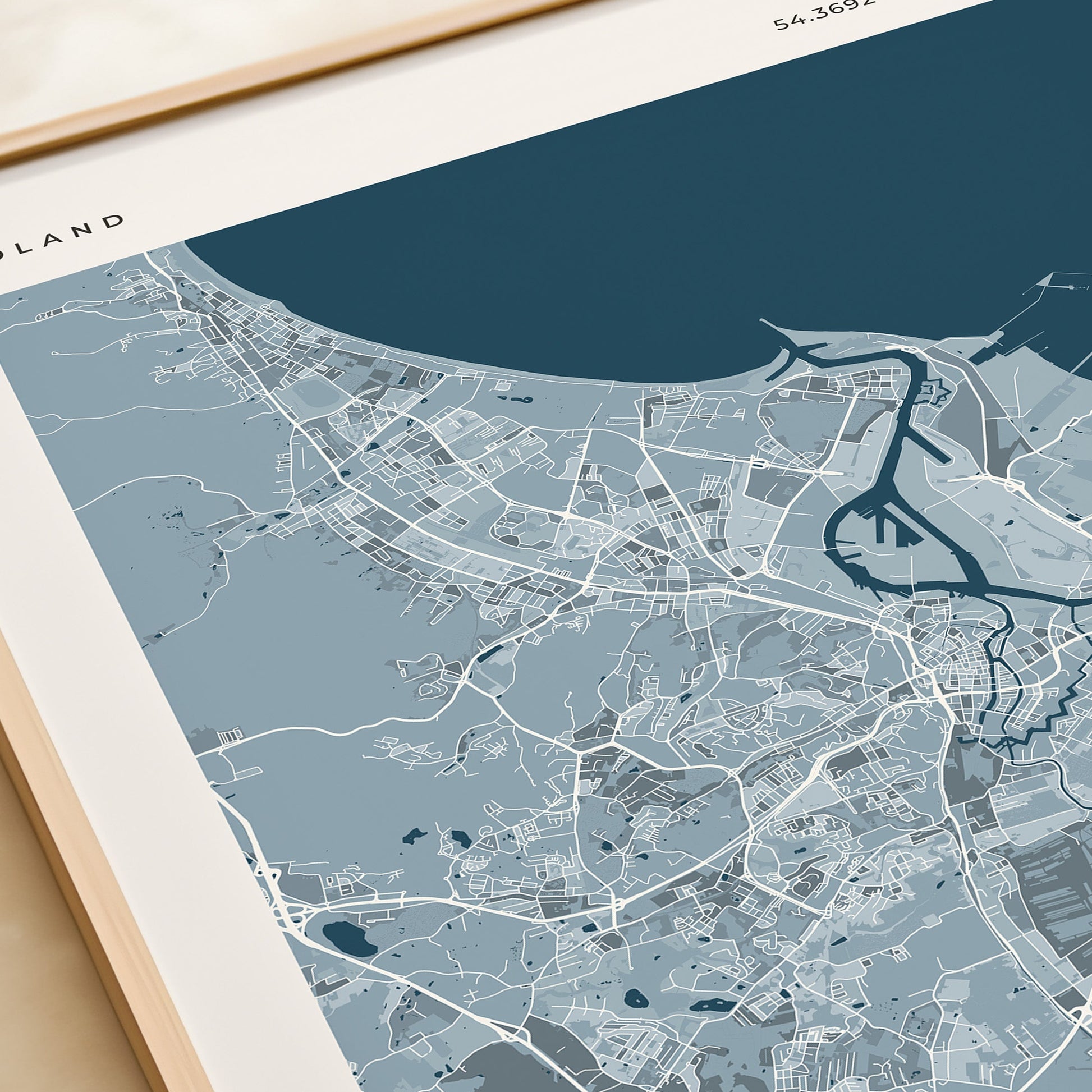 A framed minimalist city map print of Gdansk, Poland, featuring a light blue and navy street-and-coastal layout on a cream background, styled on a tabletop next to a beige gift box.