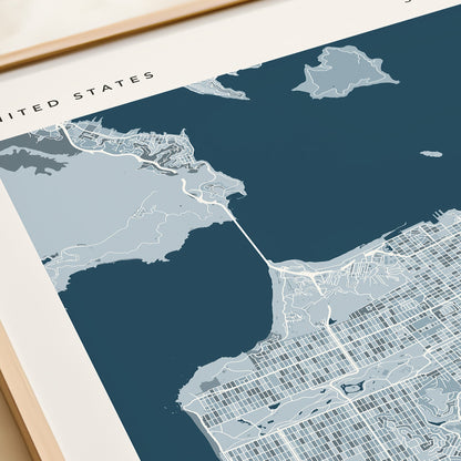 A framed San Francisco city map print with a navy blue background and light gray street details, displayed on a beige tabletop next to a rolled cream document.