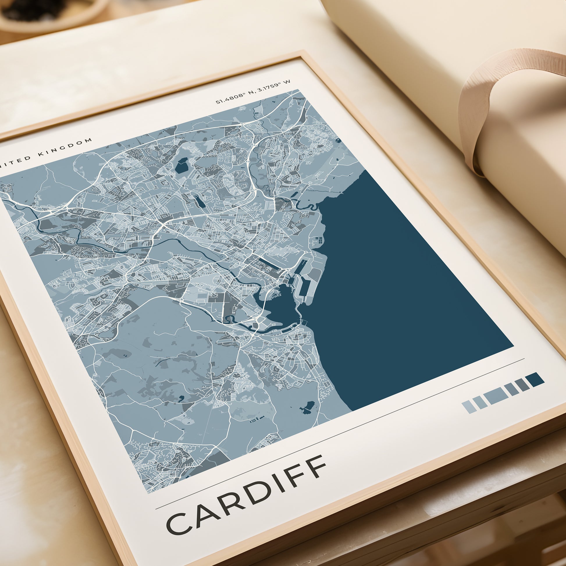 A framed minimalist map print of Cardiff, United Kingdom, featuring a modern blue and gray color palette, styled on a light beige tabletop next to a cream-colored gift box.