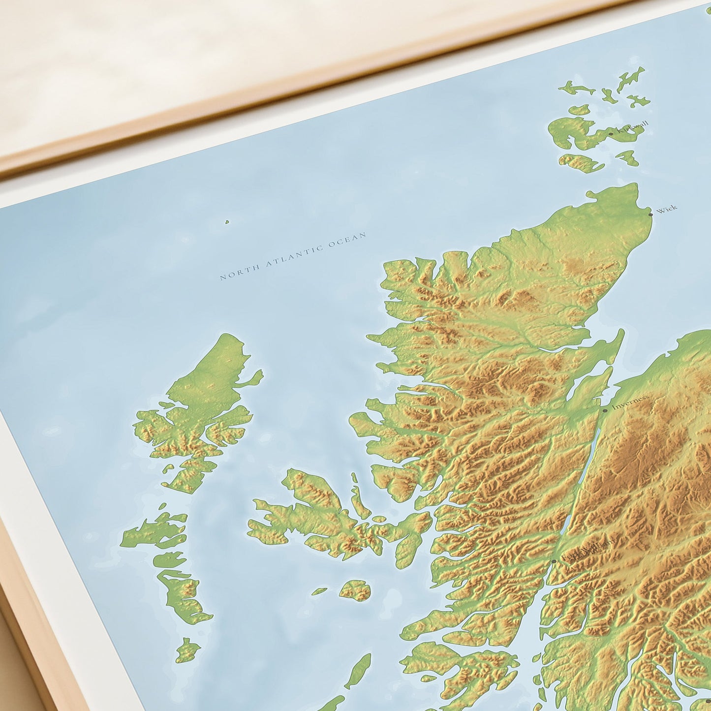 A framed Scotland topographic map print with a light blue sea area, green and yellow landmass, and the word 'SCOTLAND' at the bottom, photographed on a beige tabletop next to a rolled cream document.