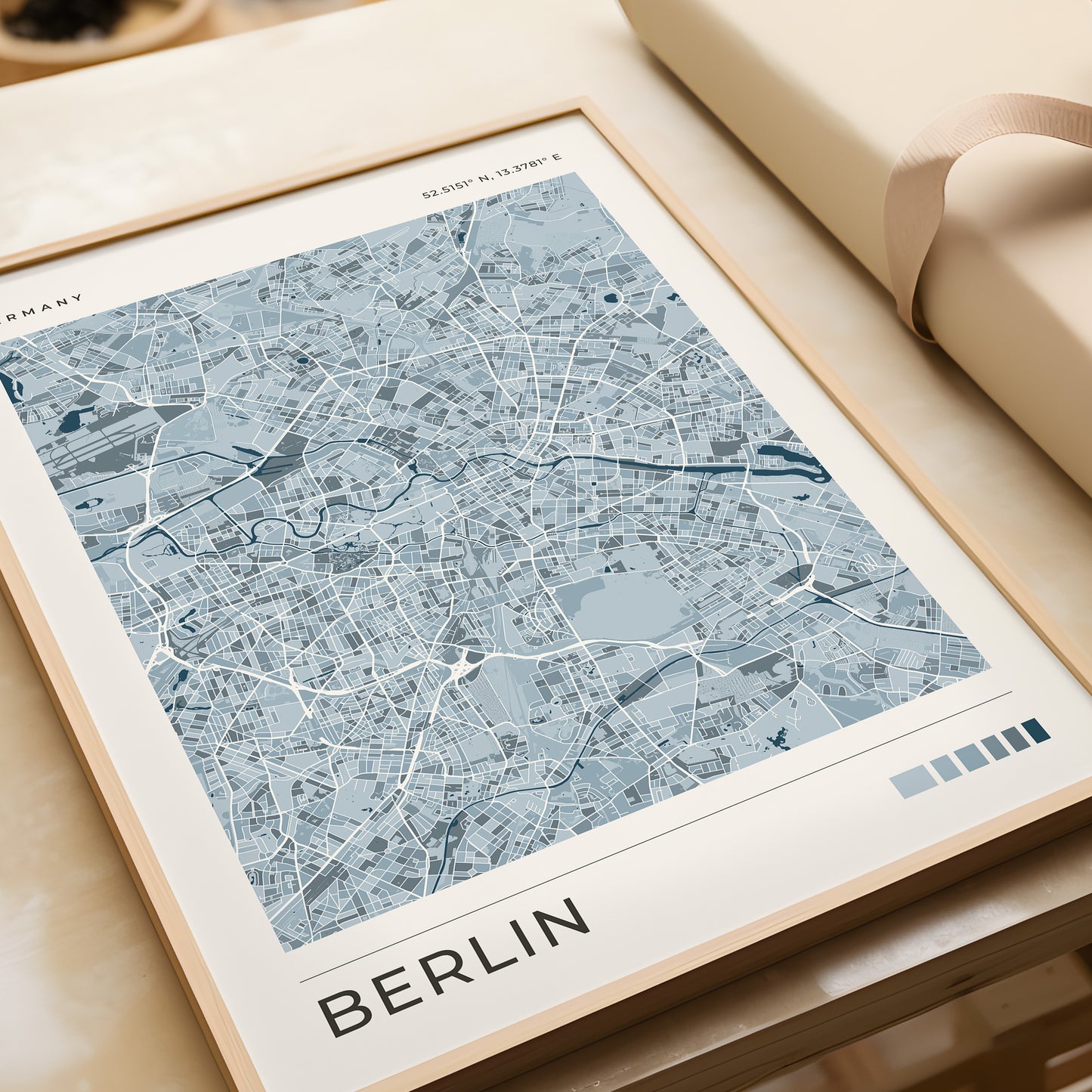 A framed city map print of Berlin, Germany, featuring a light blue and white street layout with the word 'BERLIN' at the bottom, styled on a beige surface next to a rolled cream fabric or notebook.
