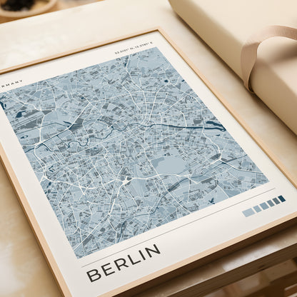 A framed city map print of Berlin, Germany, featuring a light blue and white street layout with the word 'BERLIN' at the bottom, styled on a beige surface next to a rolled cream fabric or notebook.