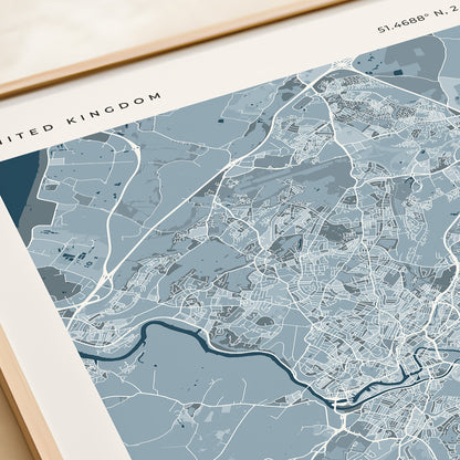 A framed city map print of Bristol, United Kingdom, featuring a light blue and gray street layout with the word BRISTOL at the bottom, styled on a beige surface next to a rolled cream paper or notebook.