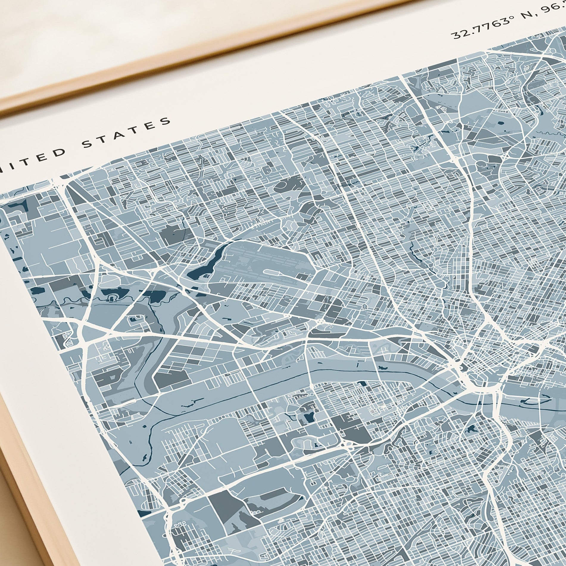 A framed modern city map print of the United States, featuring a detailed street grid and waterways in shades of slate blue, gray, and white, with a light wood frame.