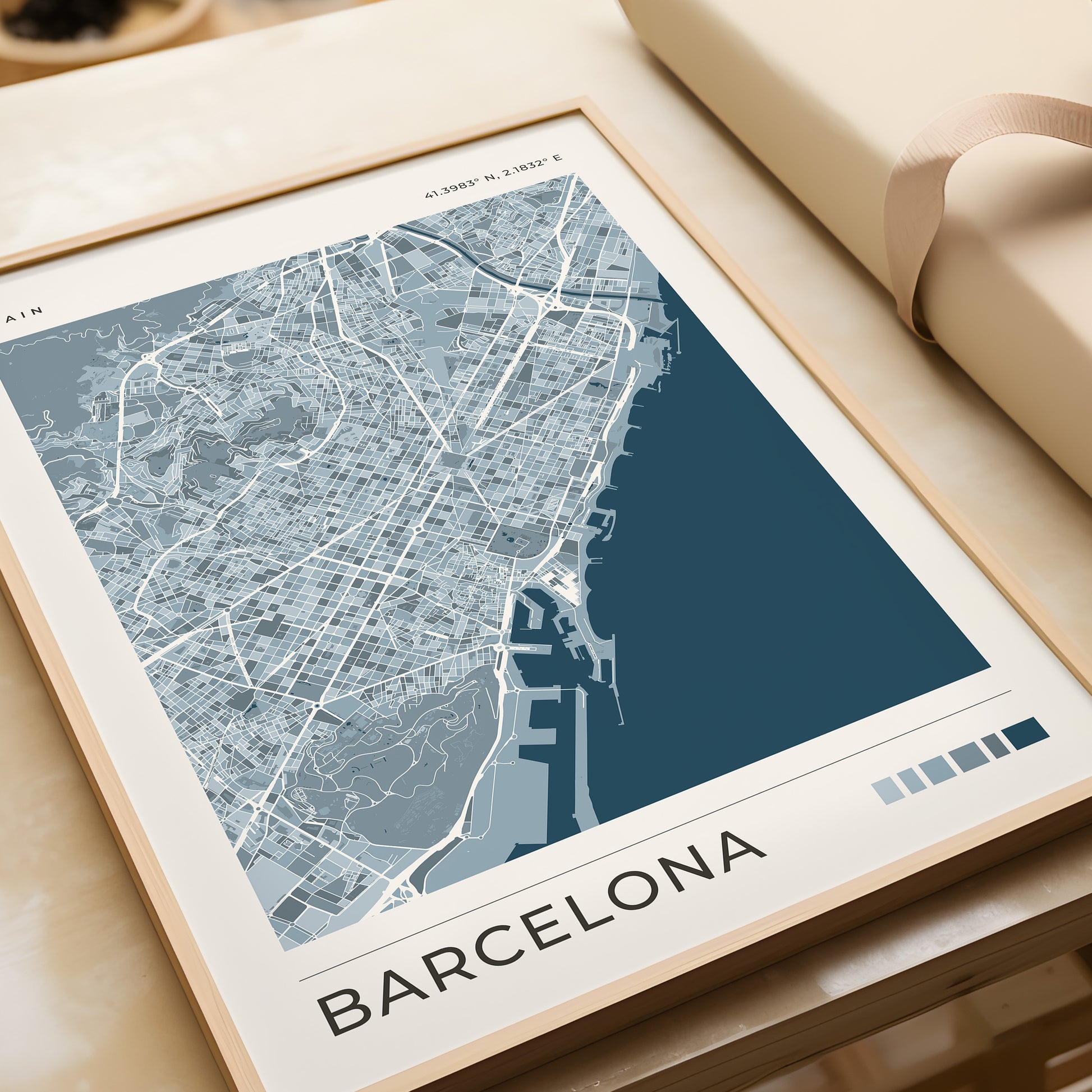 A framed Barcelona city map print with a modern street-style design in shades of blue, gray, and white, displayed on a beige tabletop next to a rolled cream document.