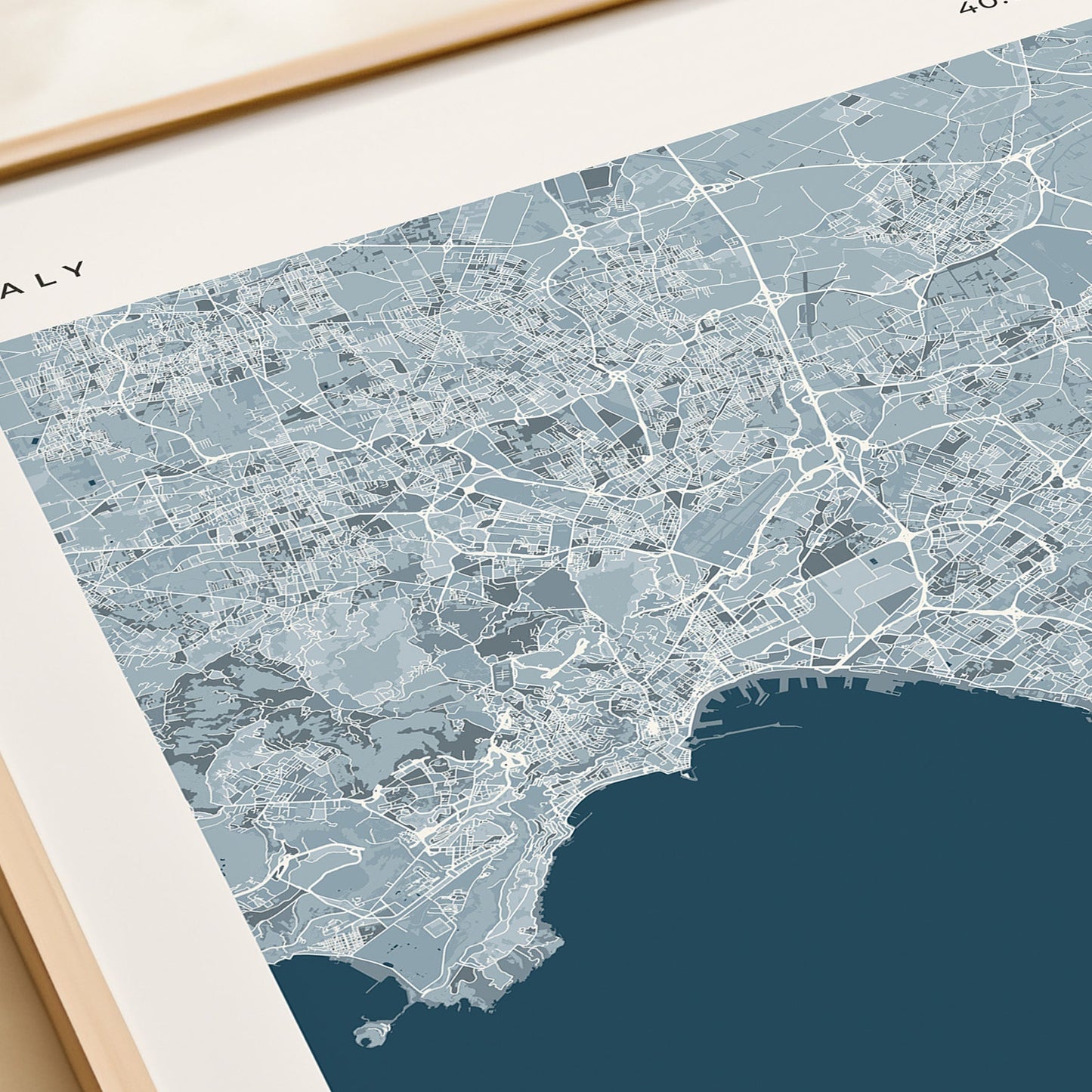 A framed Naples city map print with a cream border and a large dark blue sea area, styled on a tabletop next to a beige gift box.