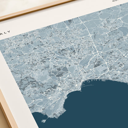 A framed Naples city map print with a cream border and a large dark blue sea area, styled on a tabletop next to a beige gift box.