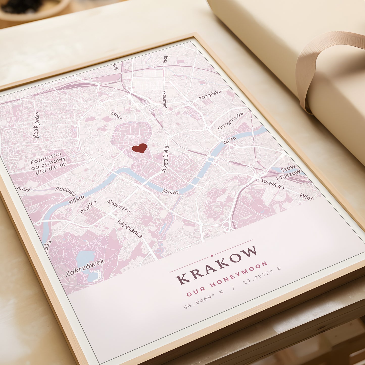 A framed personalized map print of Krakow, Poland, featuring a light pink and white street layout with a small red heart in the center and the text 'KRAKOW OUR HONEYMOON' at the bottom. The print is displayed on a light beige surface next to a cream-colored gift box.