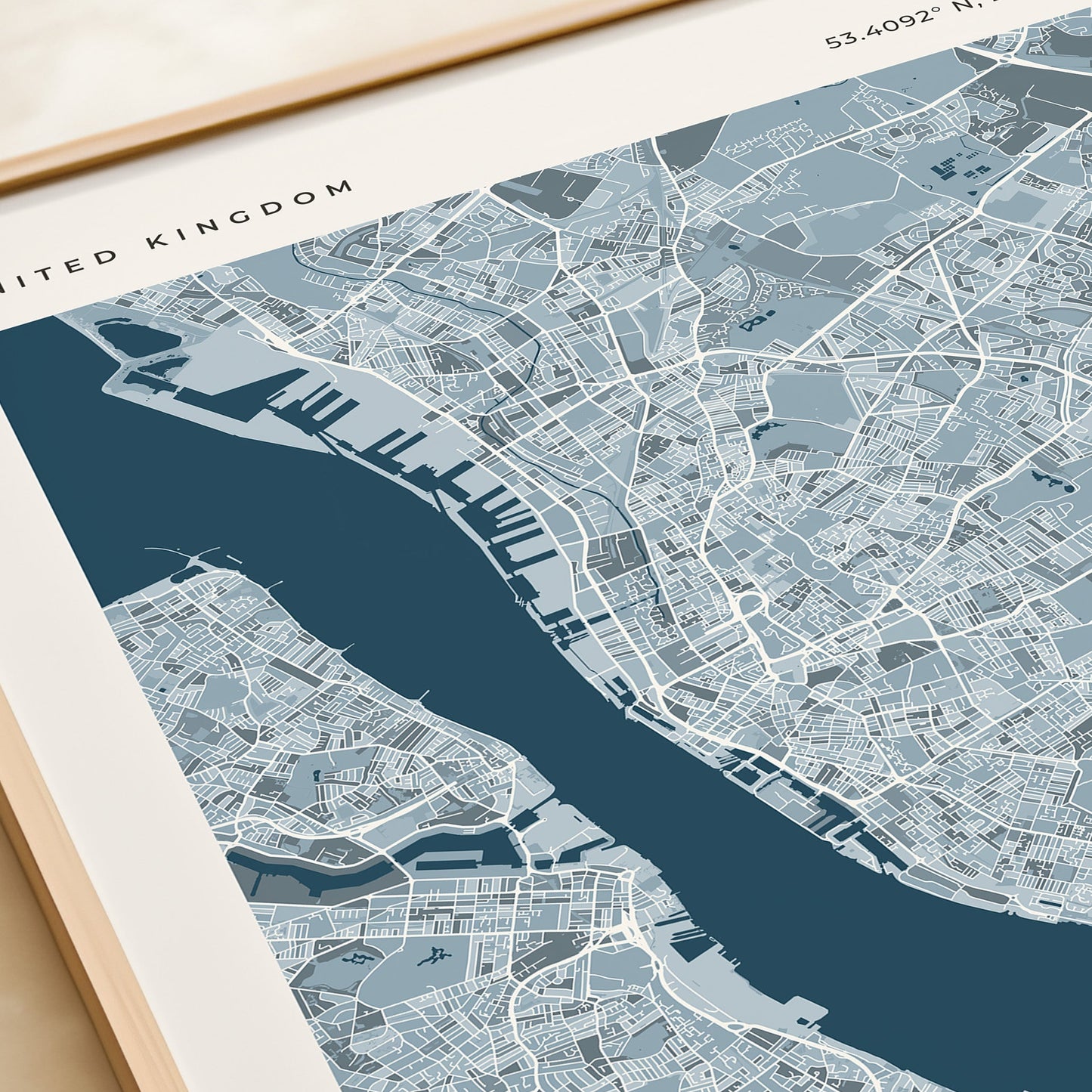 A framed map print of Liverpool, United Kingdom, featuring a modern street-style map in shades of blue and gray with a dark blue river running through the center. The print is housed in a light wood frame and styled on a beige tabletop next to a rolled cream-colored notebook or book.