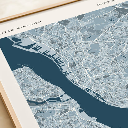 A framed map print of Liverpool, United Kingdom, featuring a modern street-style map in shades of blue and gray with a dark blue river running through the center. The print is housed in a light wood frame and styled on a beige tabletop next to a rolled cream-colored notebook or book.