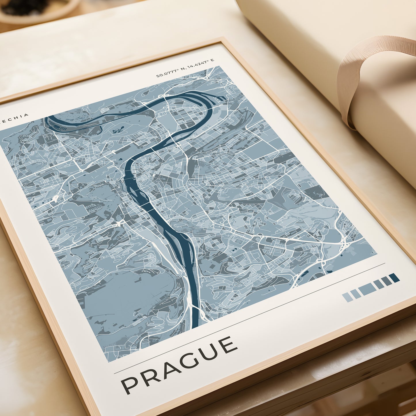 A framed city map print of Prague, shown at an angle on a light wood surface next to a beige gift box. The print features a modern map design in shades of blue, gray, and white, with the word 'PRAGUE' printed at the bottom.