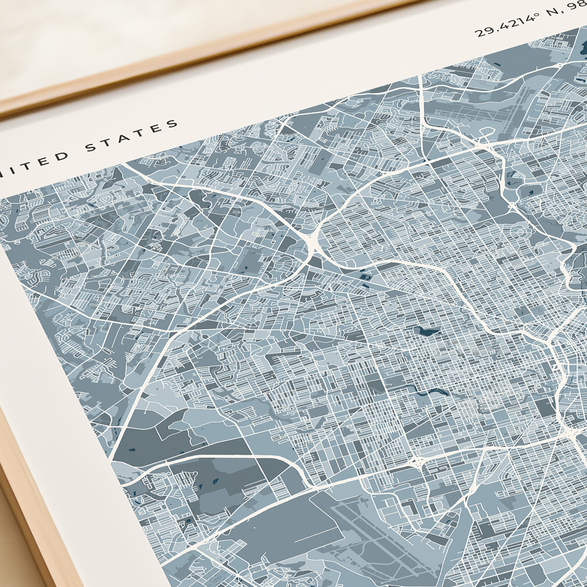 A framed city map print of the United States, featuring a detailed street grid and waterways in shades of blue, gray, and white, with a light wood frame visible.