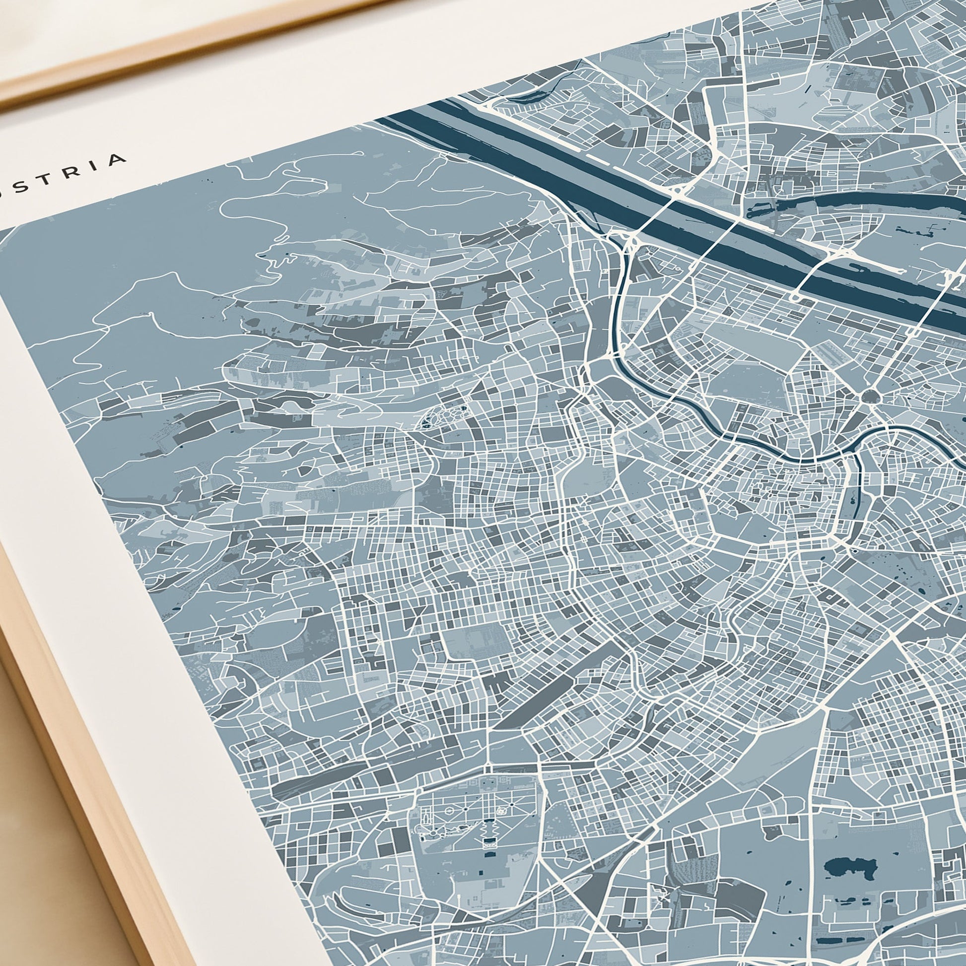 A framed minimalist map print of Vienna, Austria, featuring a light blue and white street layout with the city name 'VIENNA' at the bottom, styled on a beige surface next to a rolled cream-colored book or notebook.