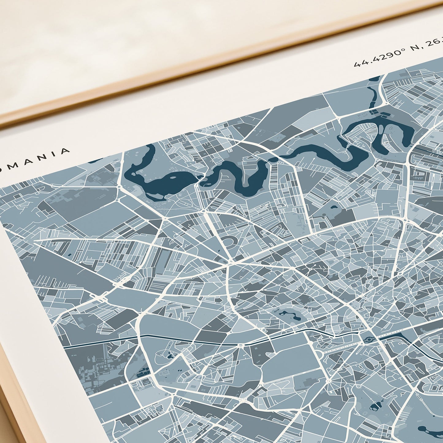 A framed city map print of Bucharest, Romania, featuring a light blue and gray street layout with dark blue text and labels, styled on a beige tabletop next to a rolled cream paper or notebook.