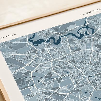 A framed city map print of Bucharest, Romania, featuring a light blue and gray street layout with dark blue text and labels, styled on a beige tabletop next to a rolled cream paper or notebook.