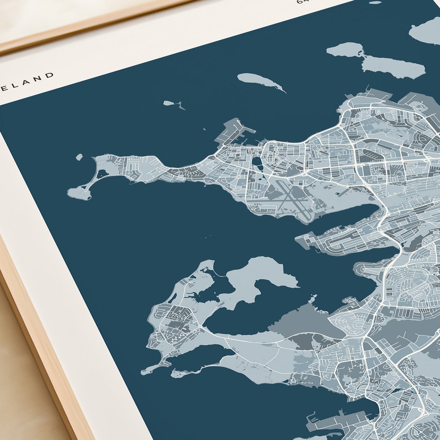 A framed city map print of Reykjavík, Iceland, shown at an angle on a light beige surface next to a cream-colored book or notebook. The print features a modern map design in shades of dark teal, light blue-gray, and white, with the text 'REYKJAVIK' printed at the bottom.