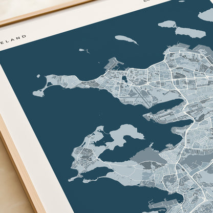 A framed city map print of Reykjavík, Iceland, shown at an angle on a light beige surface next to a cream-colored book or notebook. The print features a modern map design in shades of dark teal, light blue-gray, and white, with the text 'REYKJAVIK' printed at the bottom.