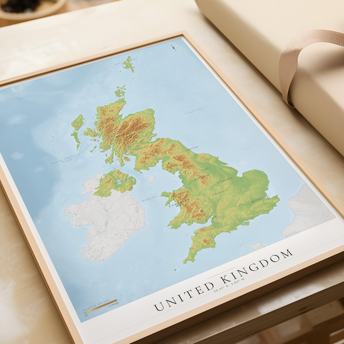 A framed topographic map poster of the United Kingdom, displayed on a light beige surface next to a rolled cream-colored book or notebook.