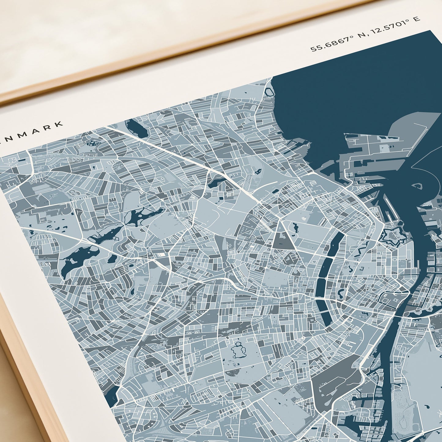 A framed Copenhagen city map print with a light wood frame, displayed on a beige tabletop next to a rolled cream paper or notebook.