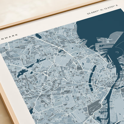 A framed Copenhagen city map print with a light wood frame, displayed on a beige tabletop next to a rolled cream paper or notebook.