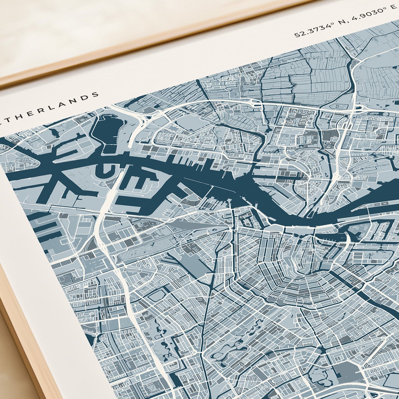 A framed Amsterdam city map print with a light wood frame, styled on a beige tabletop next to a cream-colored book or notebook.