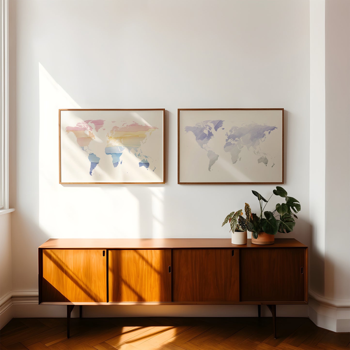 Two framed world maps with pastel watercolor colors hang on a white wall above a mid-century modern wooden sideboard with potted plants, in a sunlit room with warm sunlight and shadows.