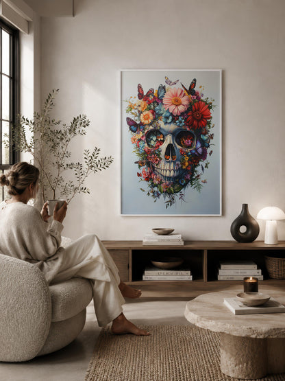 A framed colorful floral skull wall art print hangs on a beige wall in a styled living room, with a woman seated in a cream lounge chair facing the artwork. The room includes a wooden console shelf, books, a stone coffee table, and a lamp.