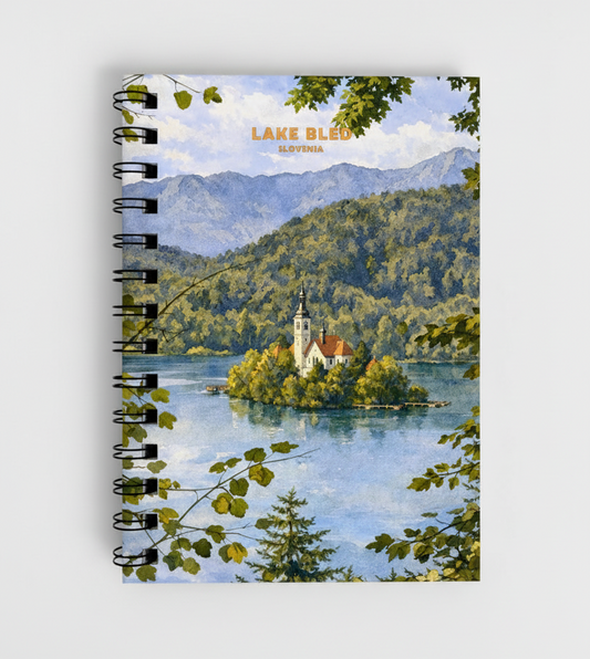 A spiral-bound notebook featuring a scenic illustration of Lake Bled, Slovenia, with a small church island, mountains, and blue water. The notebook has a black wire spiral binding and is photographed against a light gray background.
