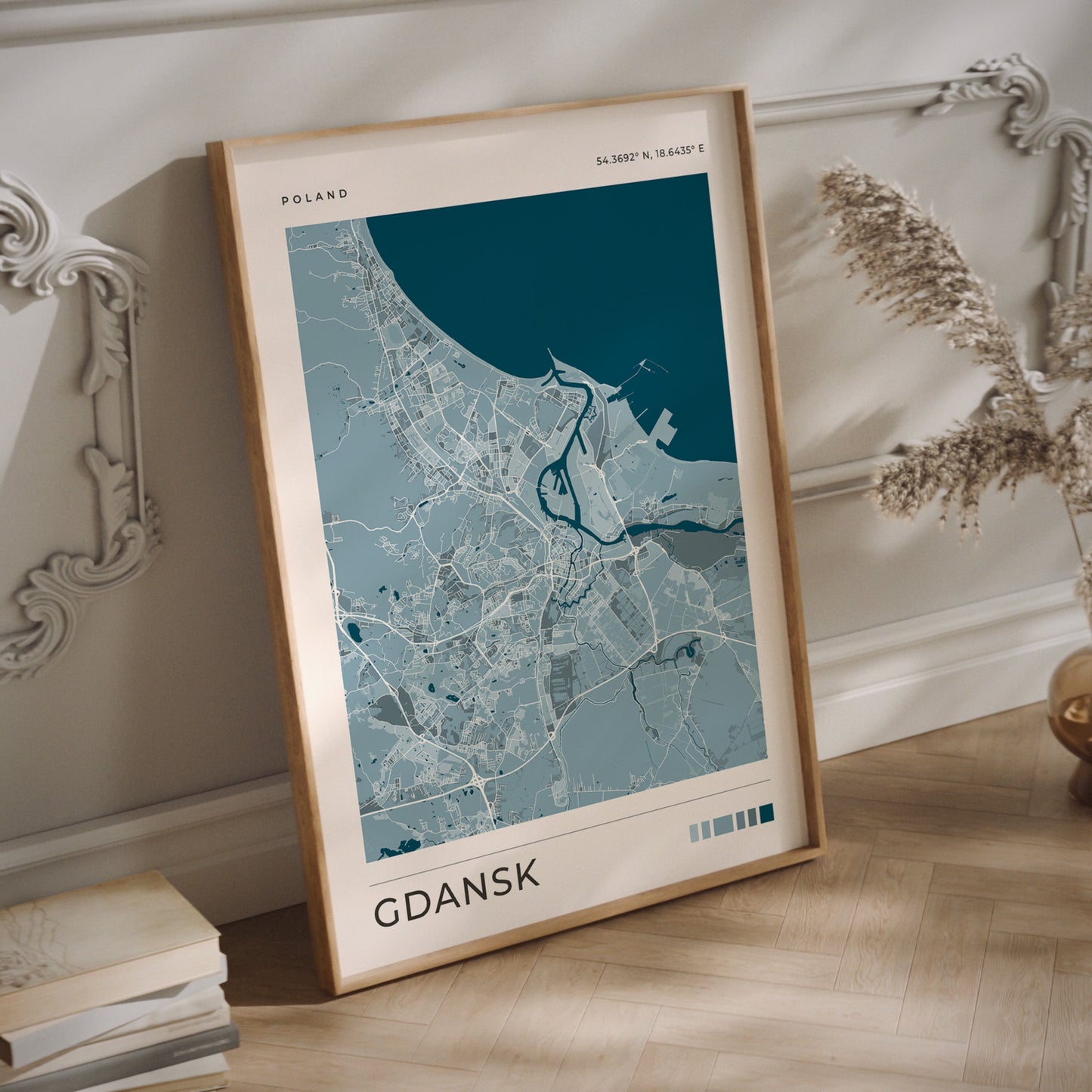 A framed city map print of Gdańsk, Poland, leaning against a white paneled wall on a light wood floor. The print features a light blue and gray map design with a dark teal water section, enclosed in a natural wood frame.