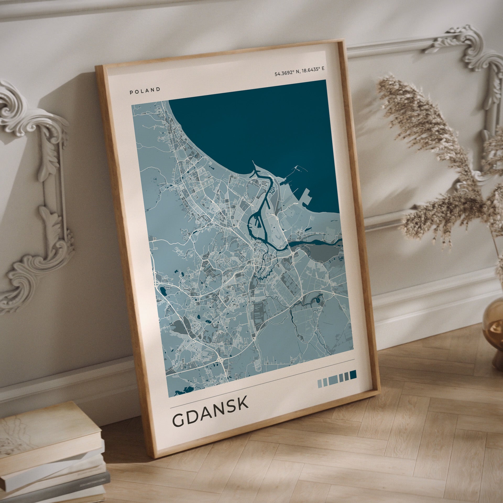 A framed city map print of Gdańsk, Poland, leaning against a white paneled wall on a light wood floor. The print features a light blue and gray map design with a dark teal water section, enclosed in a natural wood frame.