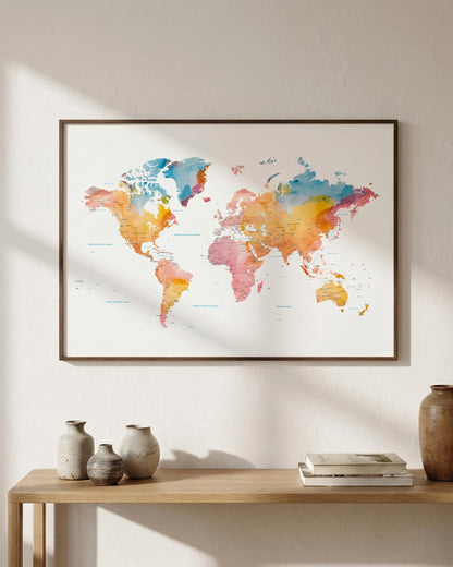 A framed world map art print with watercolor-style continents in blue, orange, pink, and yellow tones, hanging on a light beige wall above a wooden console table with decorative vases and books.