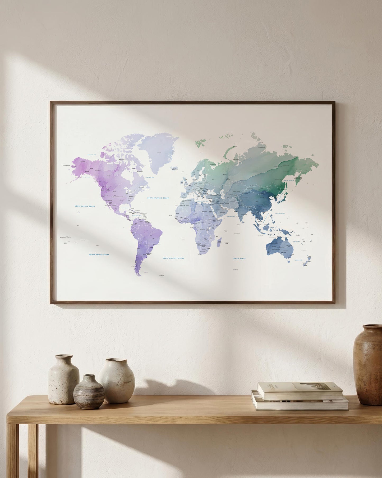 A framed world map print with a watercolor finish in soft purple, blue, and green tones, hanging on a light beige wall above a wooden console table with decorative vases and books.