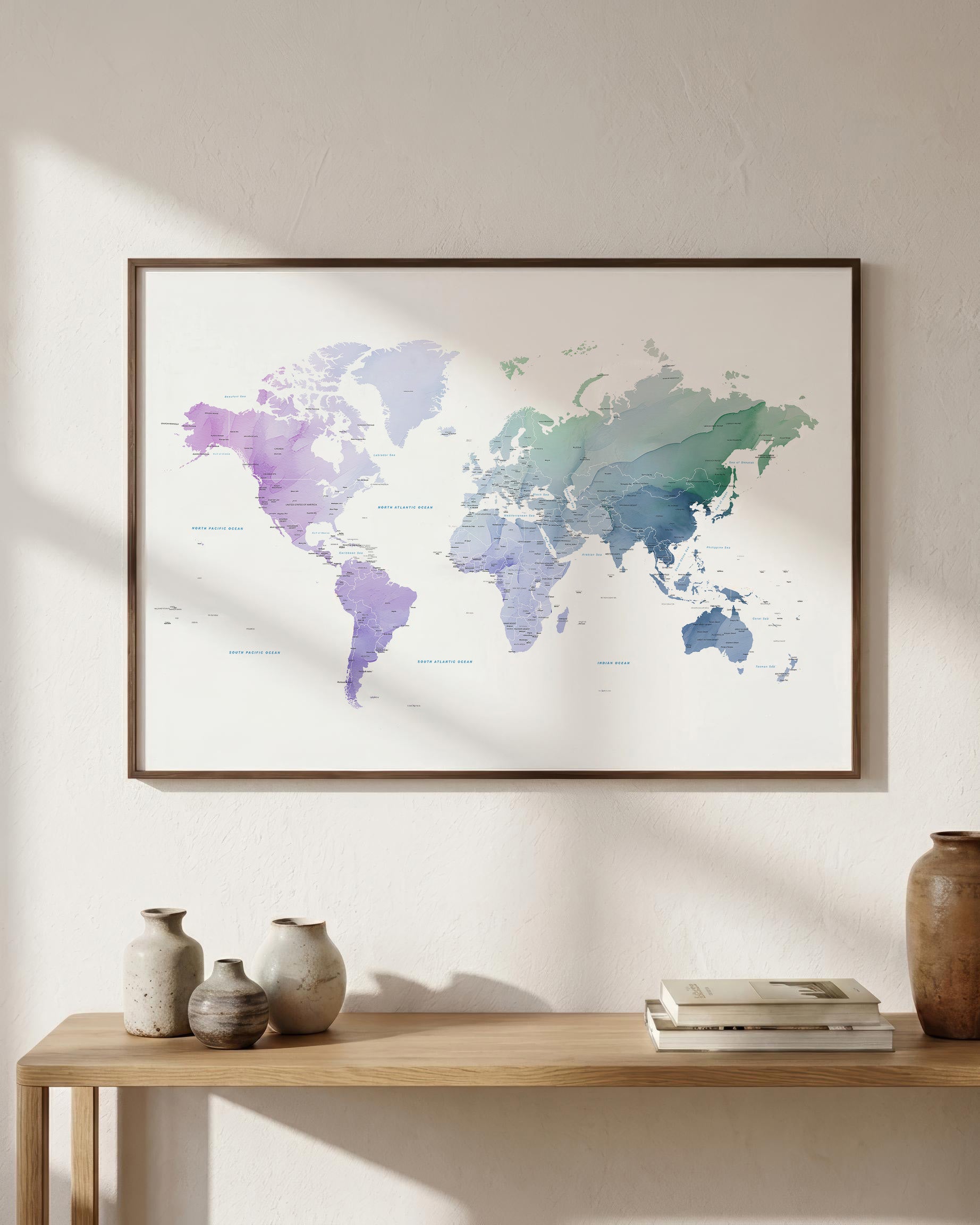 A framed world map print with a watercolor finish in soft purple, blue, and green tones, hanging on a light beige wall above a wooden console table with decorative vases and books.