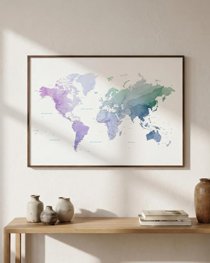 A framed world map print with a watercolor finish in soft purple, blue, and green tones, hanging on a light beige wall above a wooden console table with decorative vases and books.