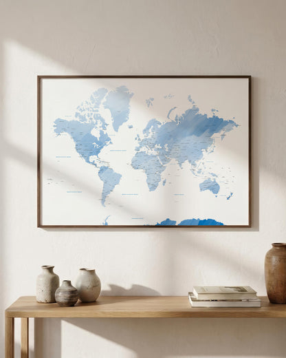 A framed world map print in light blue and white tones hangs on a beige wall above a wooden console table styled with ceramic vases and books.