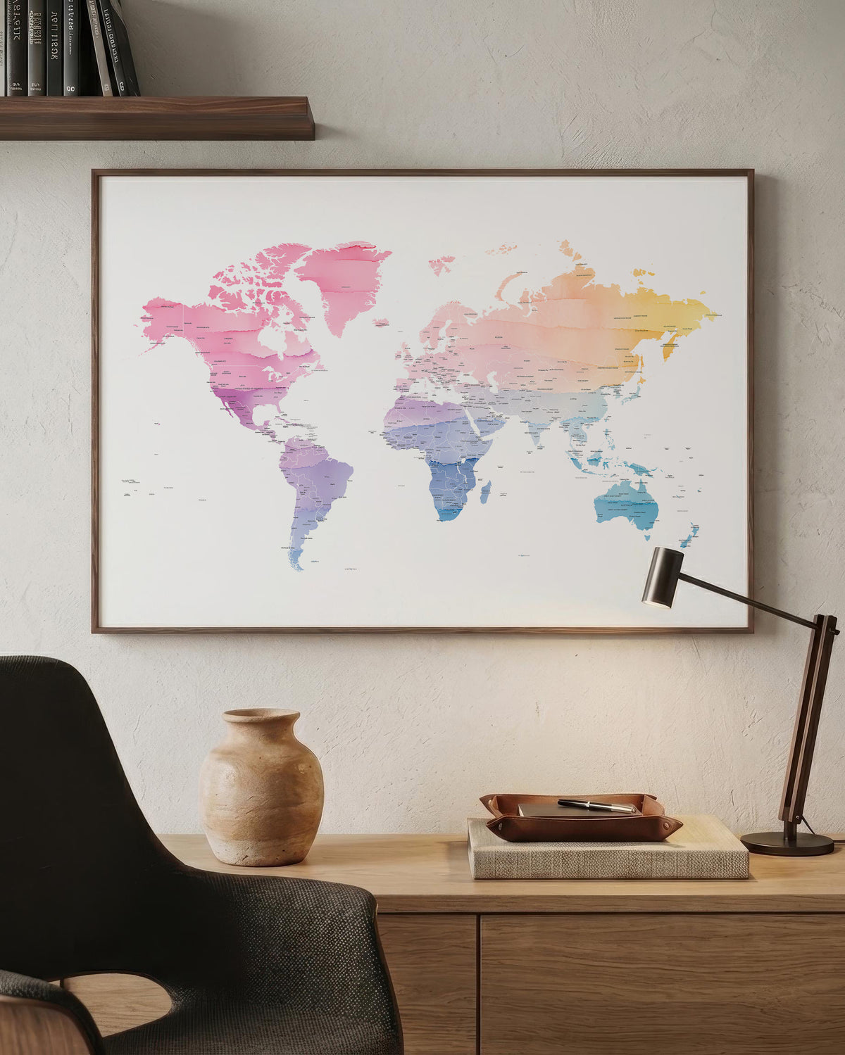 A framed world map poster with a watercolor design in pink, purple, blue, and yellow tones, hanging on a textured beige wall above a wooden desk with a lamp, vase, and chair.