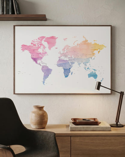 A framed world map poster with a watercolor design in pink, purple, blue, and yellow tones, hanging on a textured beige wall above a wooden desk with a lamp, vase, and chair.