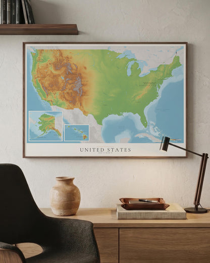 A framed United States topographic map print hangs on a textured off-white wall above a wooden desk with a black chair, ceramic vase, and desk lamp.