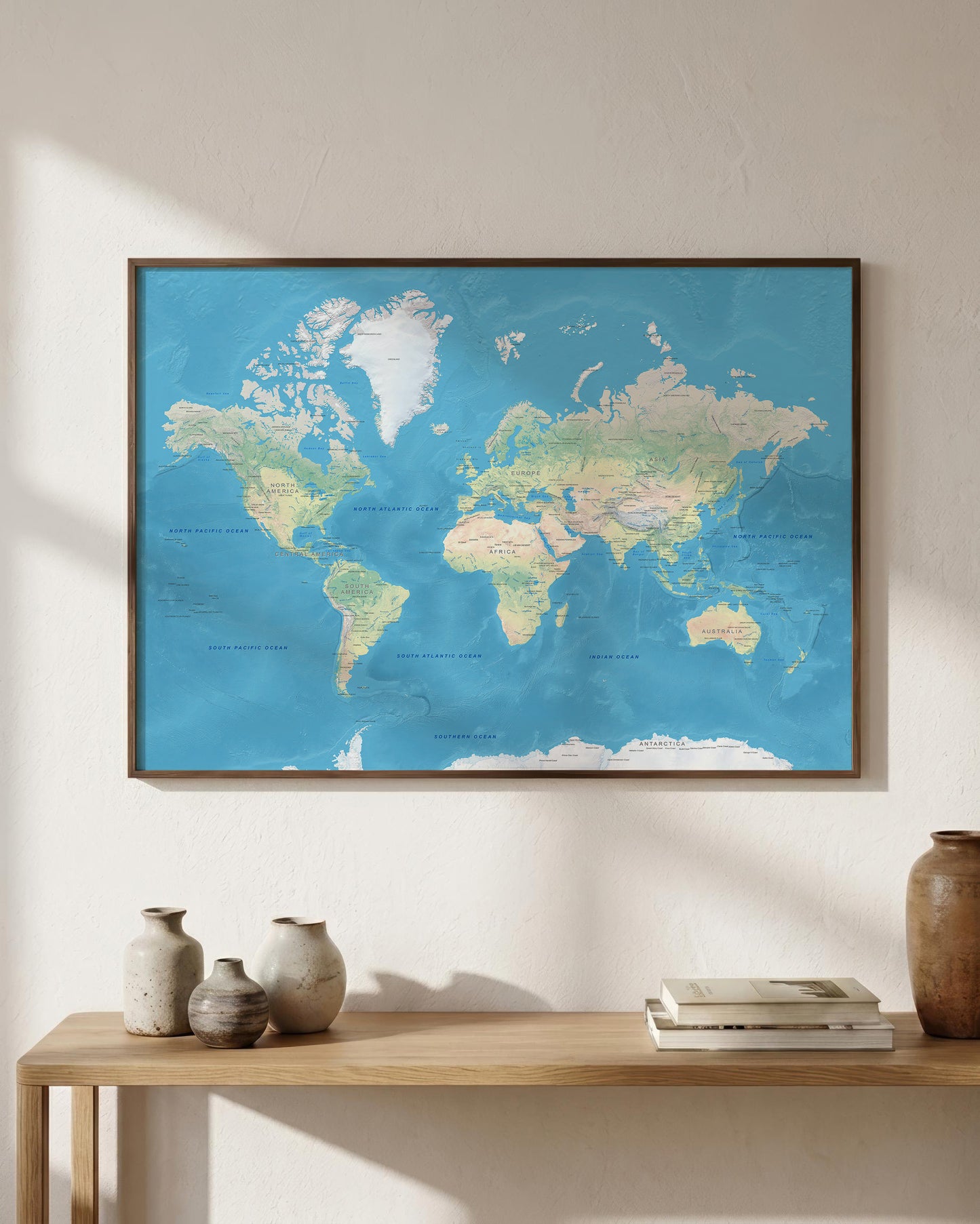 A framed world map poster with a blue ocean and beige landmasses, displayed on a light beige wall above a wooden console table with decorative vases and books.