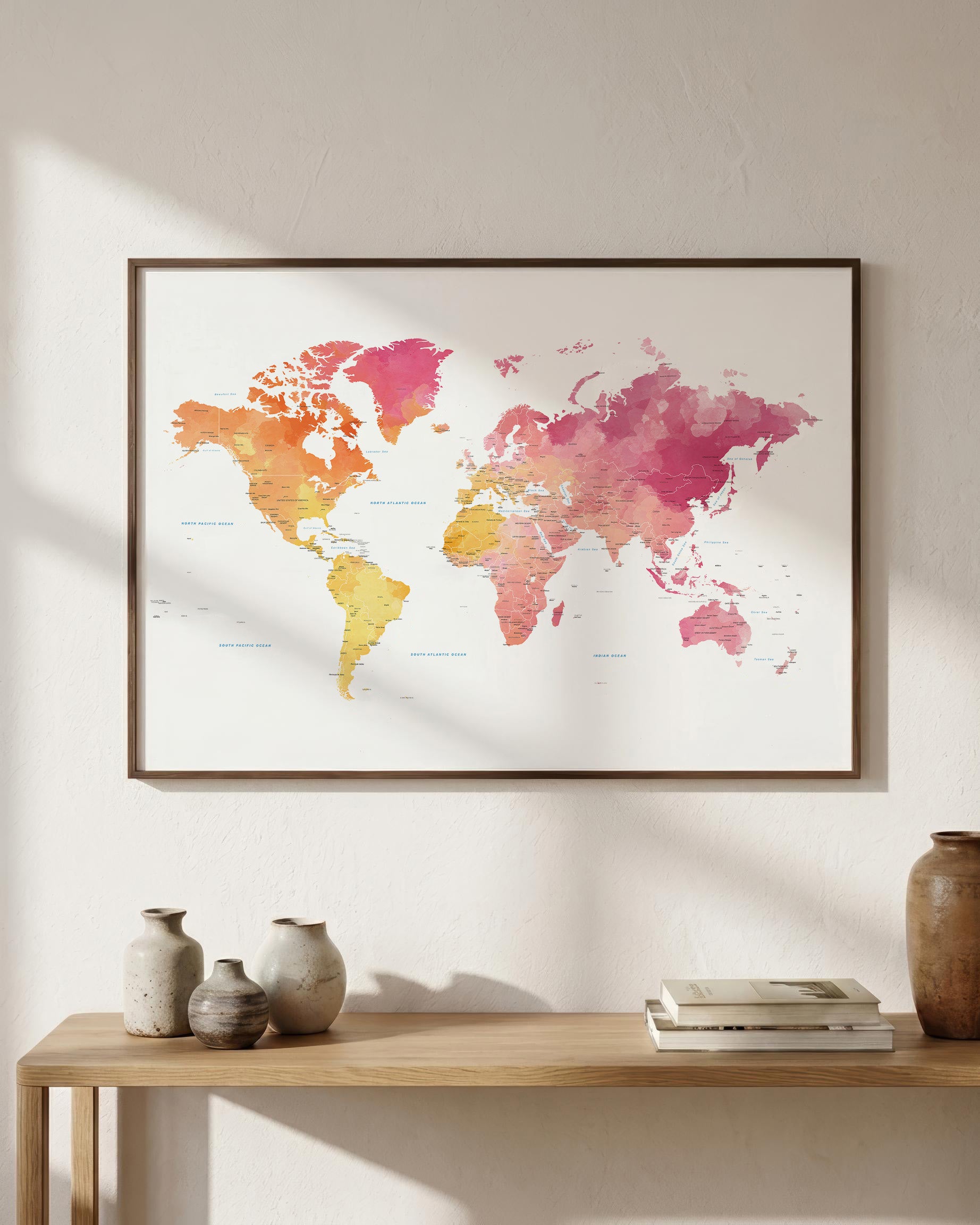 A framed world map watercolor print in pink, orange, and yellow tones, displayed on a light beige wall above a wooden console table with decorative vases and books.