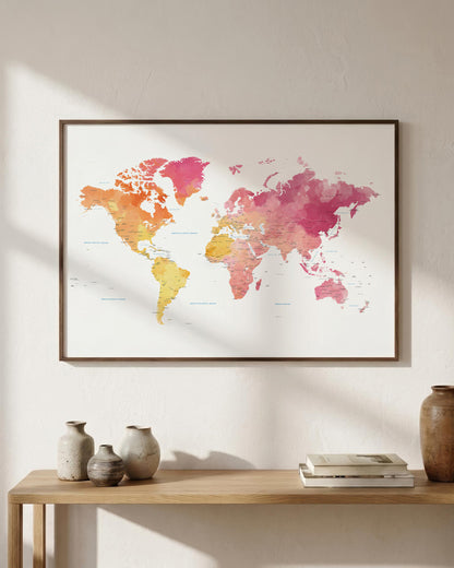 A framed world map watercolor print in pink, orange, and yellow tones, displayed on a light beige wall above a wooden console table with decorative vases and books.