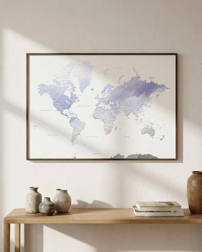 A framed world map print with soft blue and purple watercolor-style landmasses hanging on a light beige wall above a wooden console table with decorative vases and books.